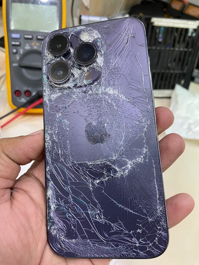 An iPhone Pro with its back glass and camera lenses shattered, held up on the bench